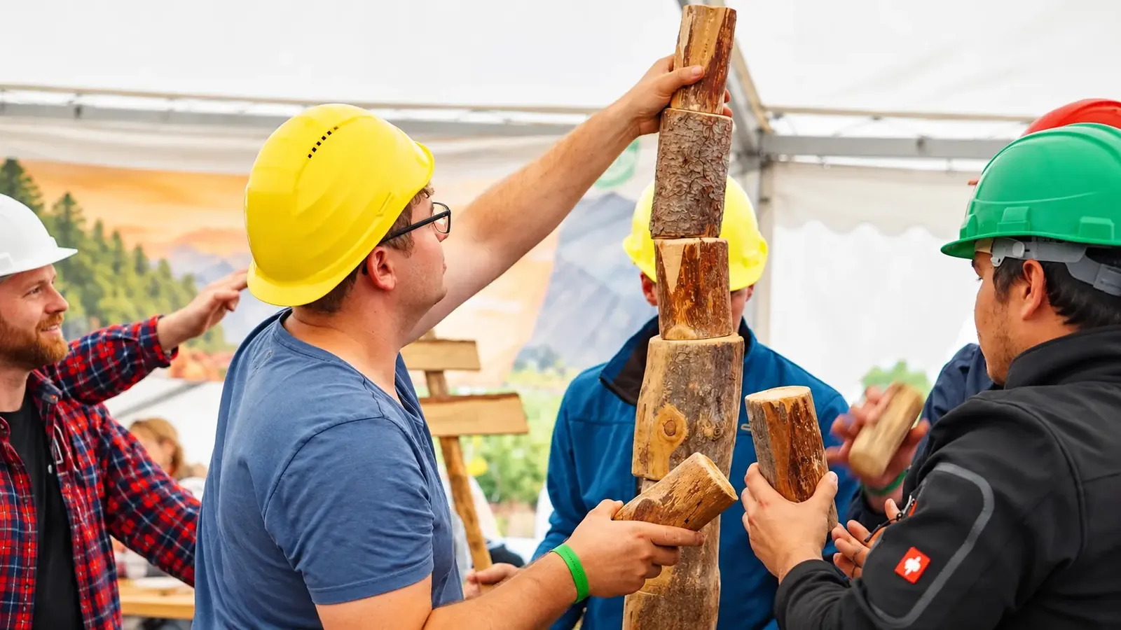 Teammitglieder mit Helmen stapeln gemeinsam Holzklötze zu einem hohen Turm im Zelt