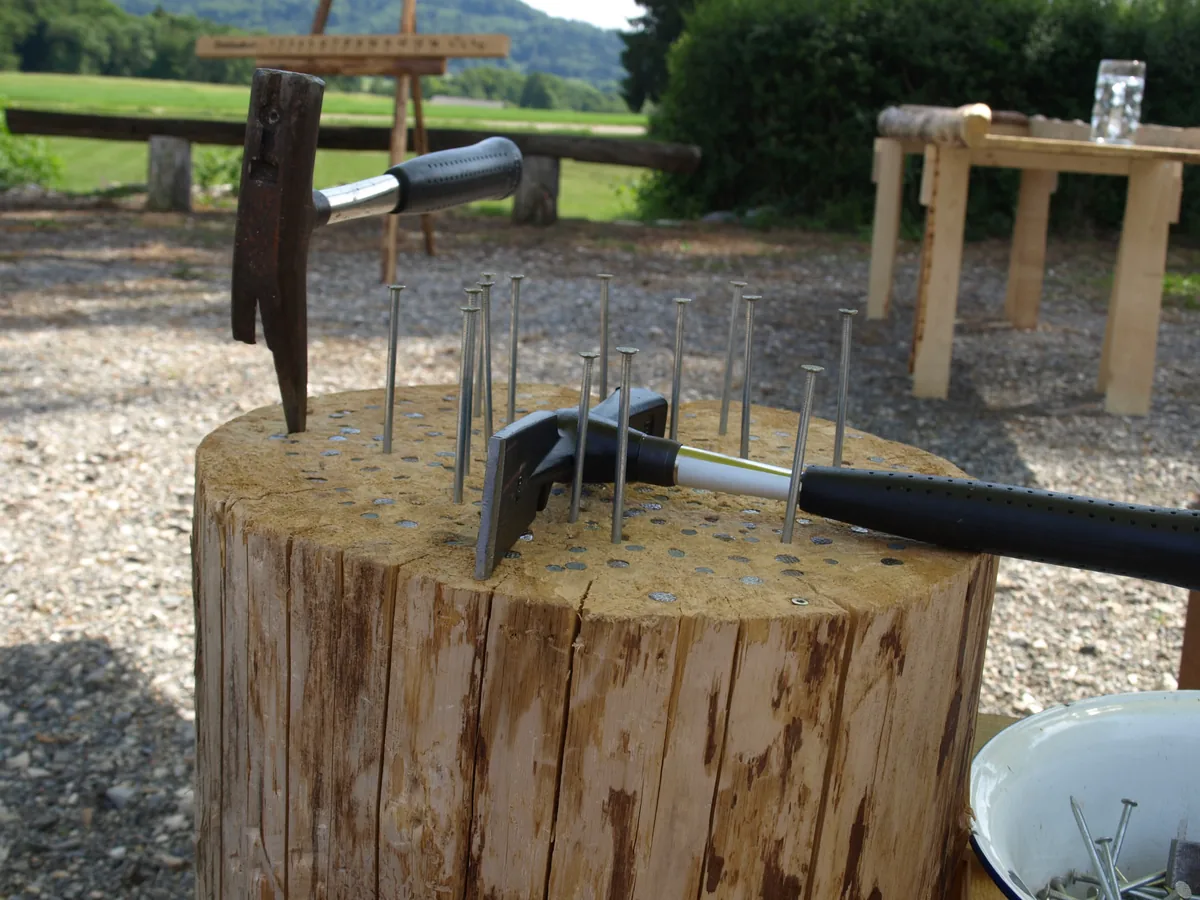 Hau den Nagel: massiver Holzstamm mit bereits eingeschlagenen Stahlnägeln, zwei Hämmern und einer Schüssel mit Nägeln, dahinter ein Punktebrett auf einer Holzbank im Freien – Holzspiel zum Mieten im Hochschwarzwald