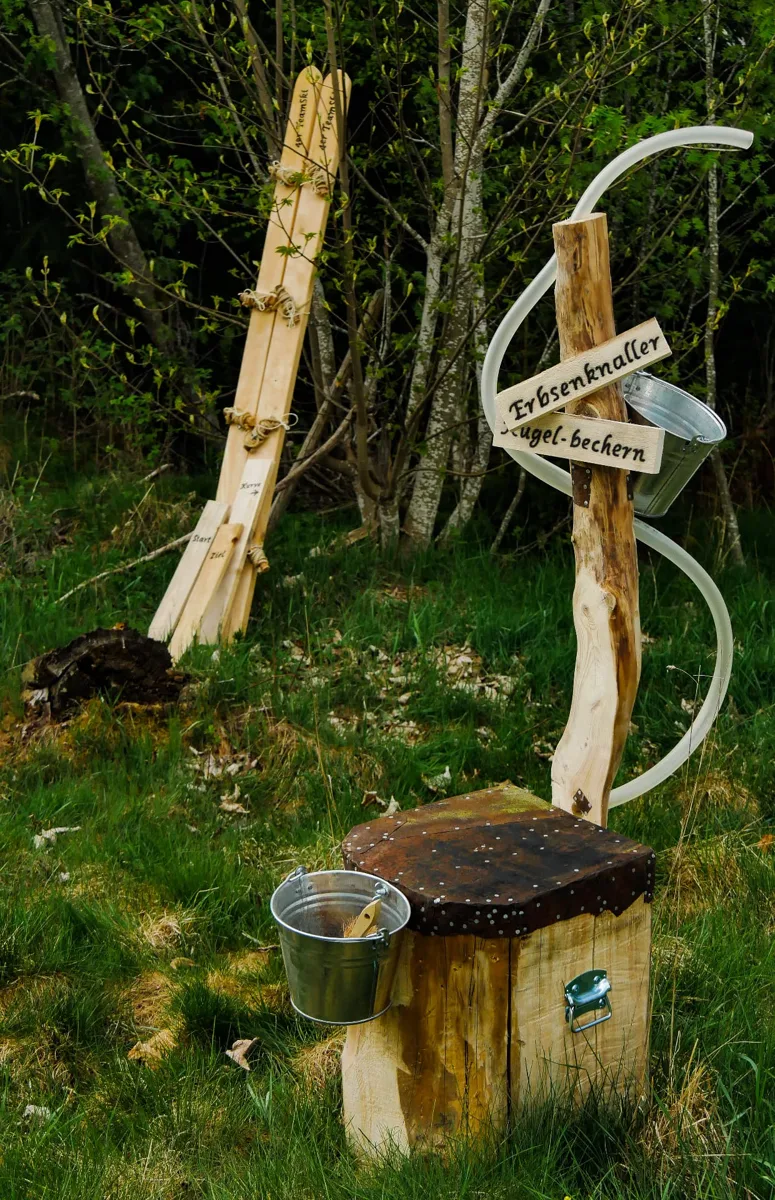 Erbsenknaller: Holzpfosten mit gewundenem Schlauch, Blecheimern und Holzklotz mit Metallfläche, Schild „Erbsenknaller“ – Holzspiel zum Mieten im Hochschwarzwald
